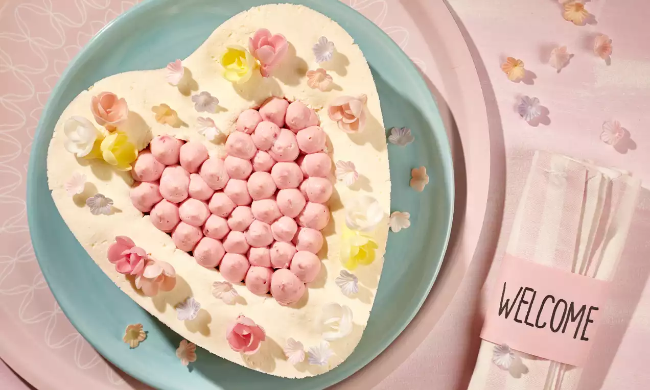 Tarte en forme de cœur garnie de crème blanche et pépites roses, décorée de fleurs délicates sur une assiette bleue