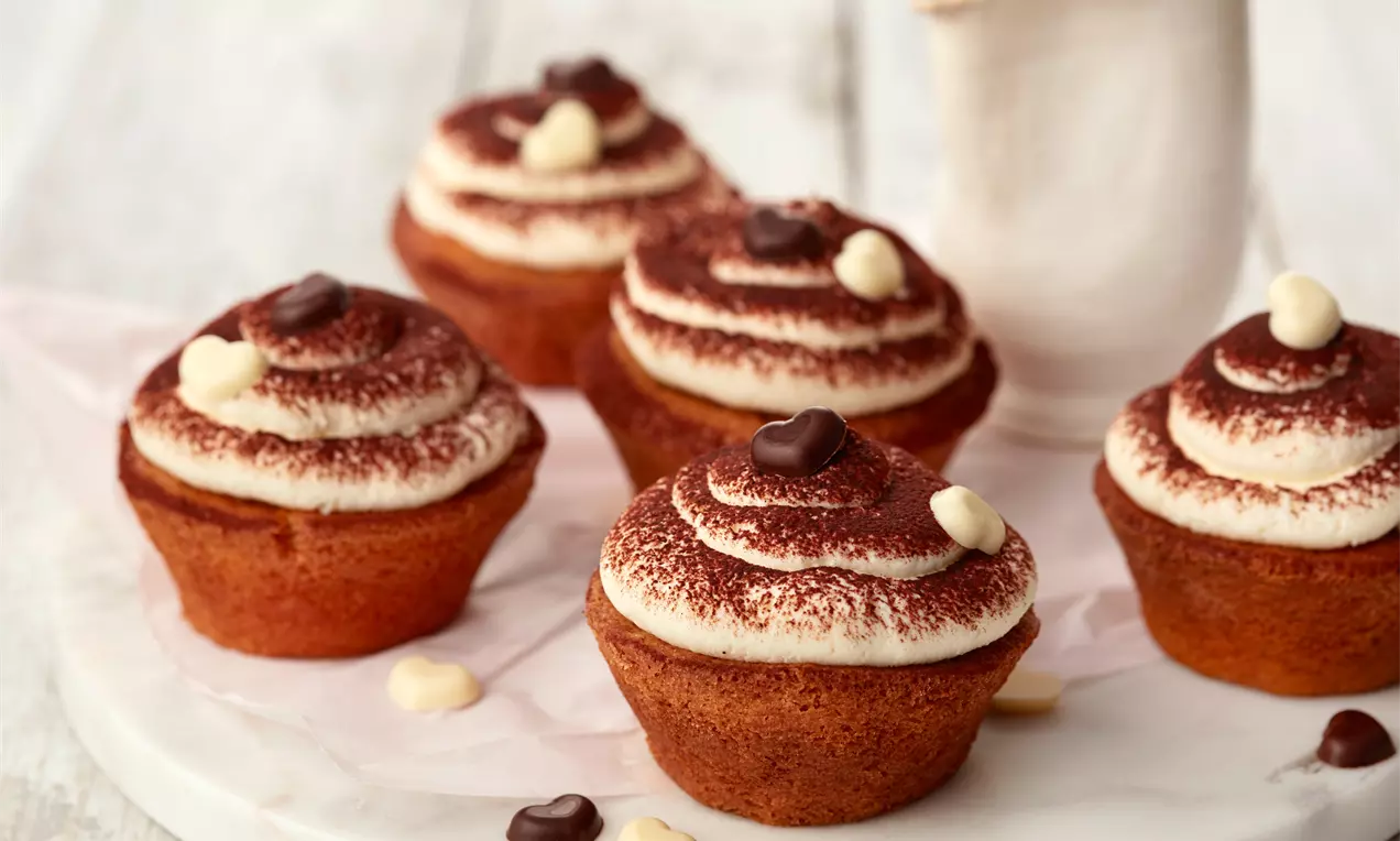 Tiramisu cupcakeovi s kremastim bijelim vrhom posutim tamnim kakao prahom, ukrašeni čokoladnim srcima na bijelom tanjiru