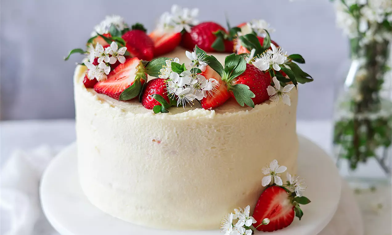 Кръгла торта с гладко бял шоколадов крем, украсена с цели и нарязани ягоди и бели цветчета, поставена върху бяла порцеланова чиния