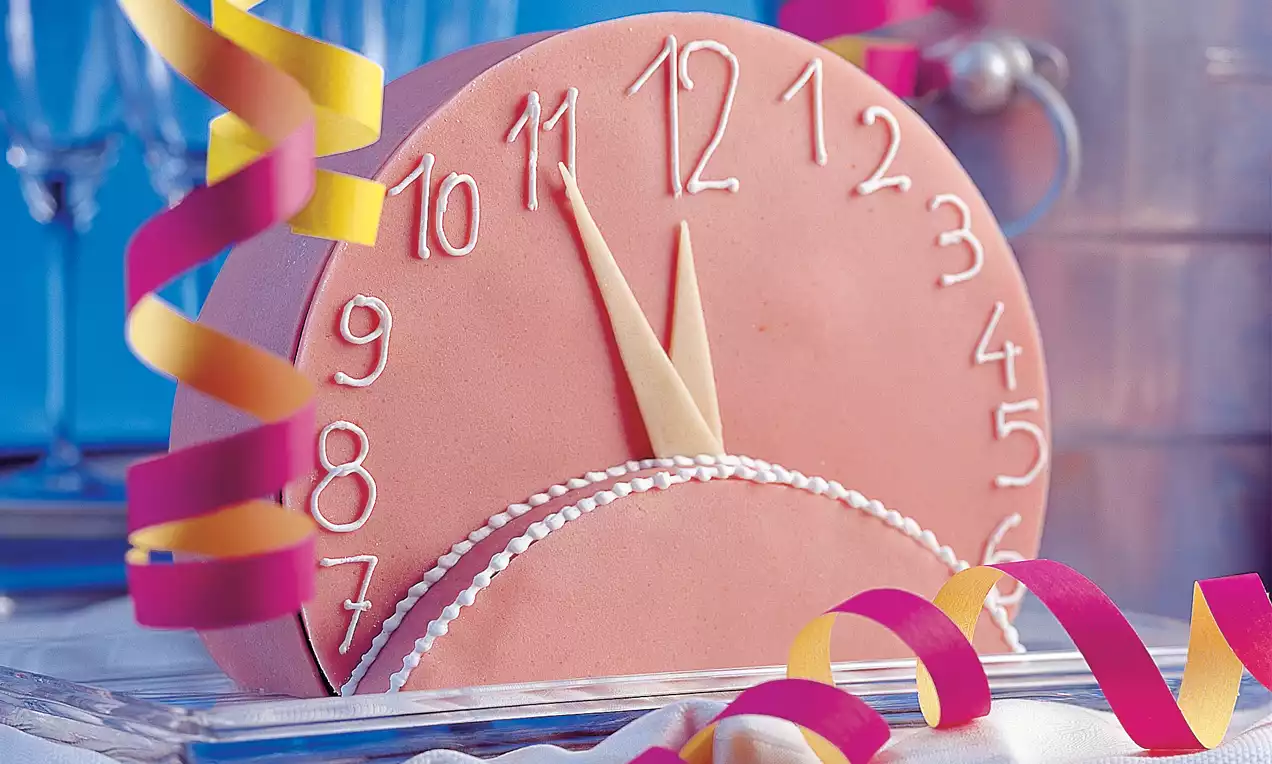 Okrugla torta boje ružičastog zlata s bijelim oznakama sata, kremastim kazaljkama i ukrasnim vrpcama na staklenom tanjuru