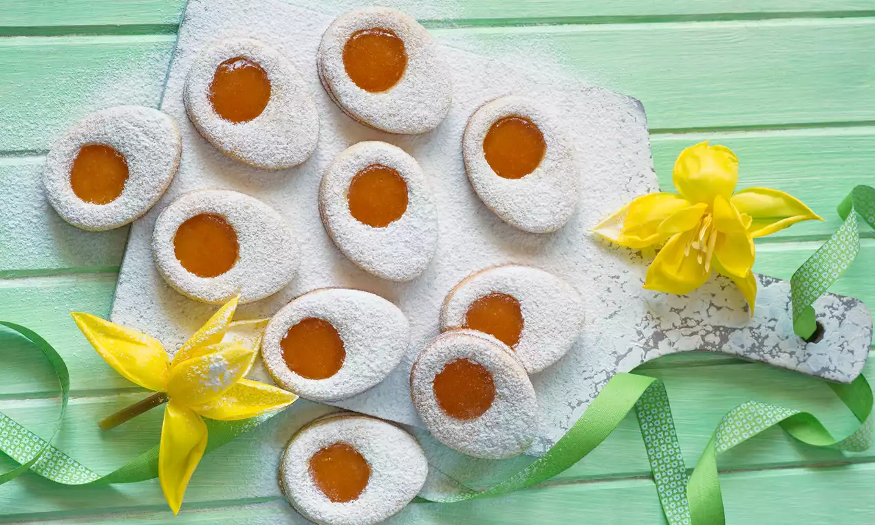 Krhki piškoti v obliki jajc z belo sladkano površino in rumeno marelično sredico, razporejeni na svetli deski z rumenimi narcismi