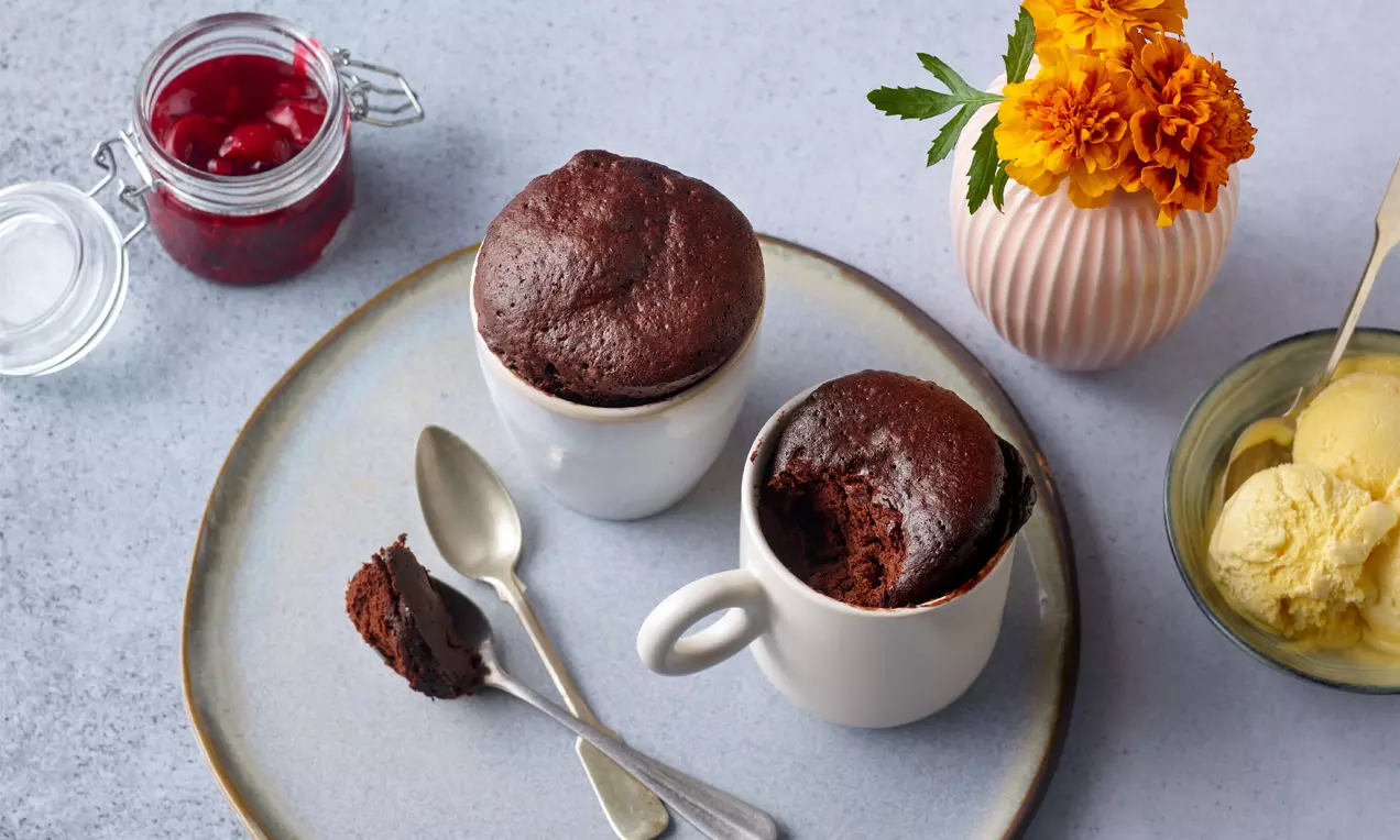 Dunkler, fluffiger Schokoladen-Tassenkuchen mit rissiger Oberfläche in weißem Porzellan auf grauem Teller, daneben Löffel, Eis und Blumenvasen
