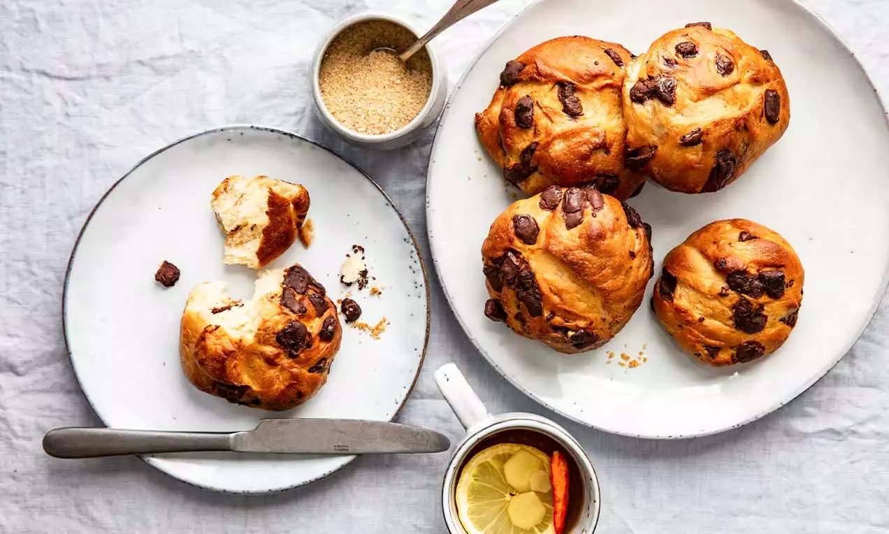 Zaobljena peciva zlatnosmeđe boje prošarana tamnim čokoladnim komadićima, na bijelim tanjurima uz šalicu čaja i šećer u posudici