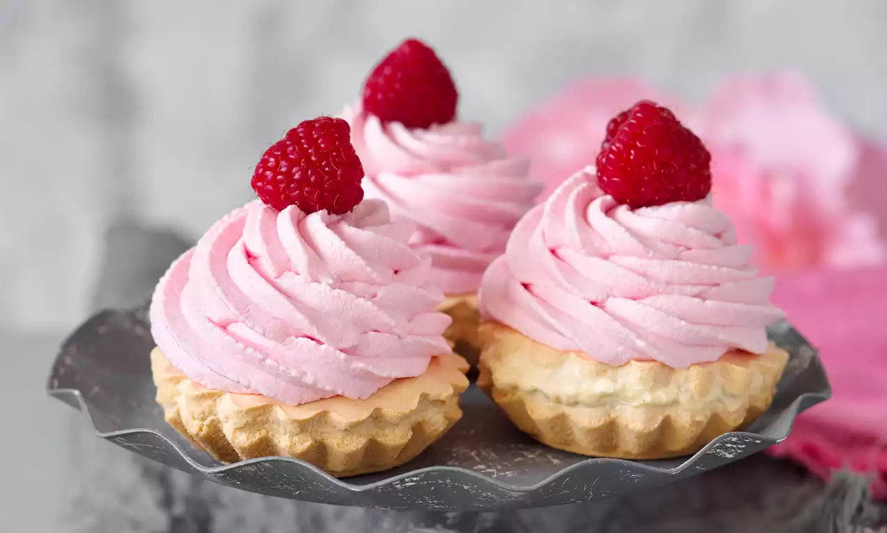 Košíčky se světle hnědým rýhovaným těstem, růžovým krémem v podobě spirál, nahoře čerstvé maliny, na kovovém stojanu