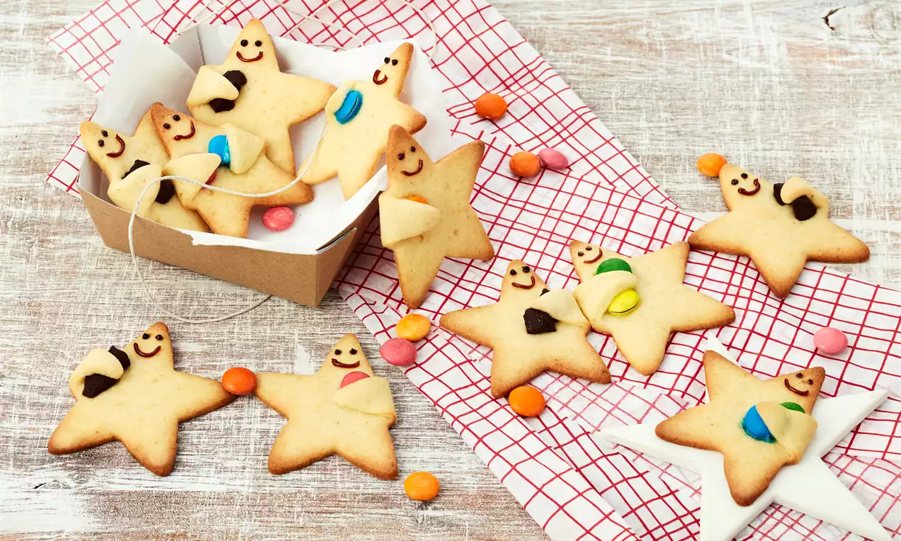 Goldene, sternförmige Plätzchen mit buntem Zuckerdekor und Schokolade, teils in einer Pappschachtel auf rot-weiß kariertem Stoff drapiert