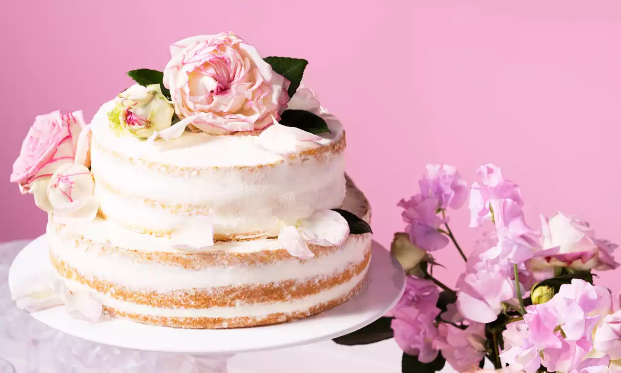 Zweistöckige Naked Cake mit weißer Creme und zarten rosa Blüten dekoriert auf einer weißen Tortenplatte vor rosa Hintergrund