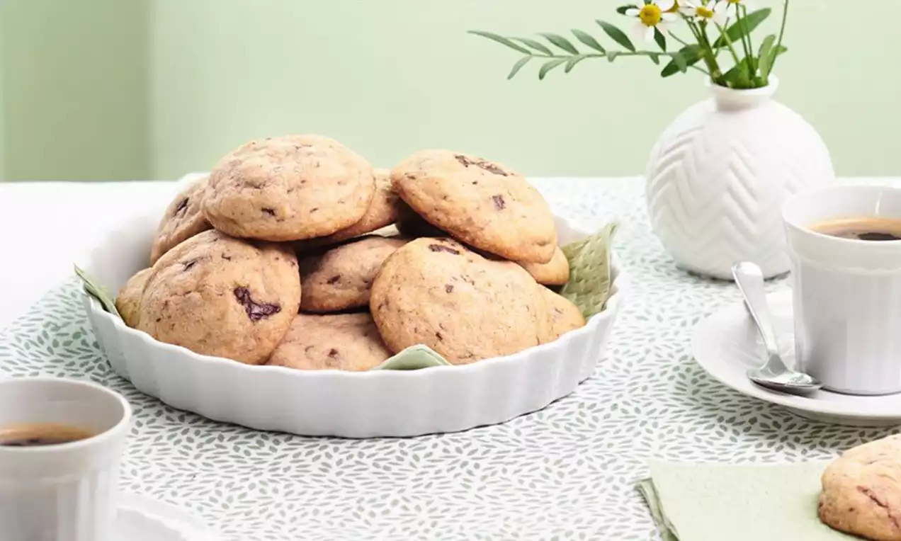Goldbraune Cookies mit Schokostückchen in einer weißen flachen Schale auf grün gemusterter Tischdecke, dekoriert mit Blumen und Kaffeetassen