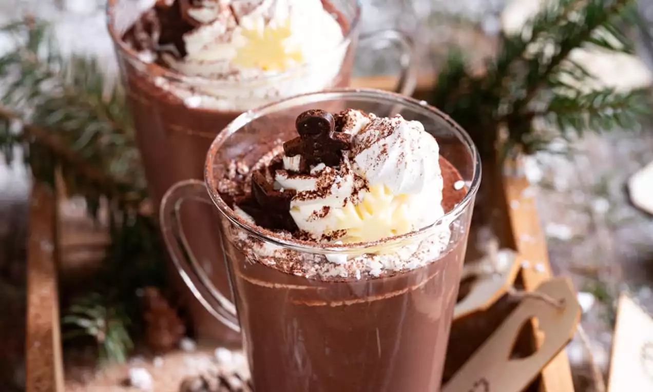 Topla čokolada globoko rjave barve v steklenem kozarcu, okrašena s puhasto belo smetano, čokoladnimi drobtinami in nežno rumenim marcipanom