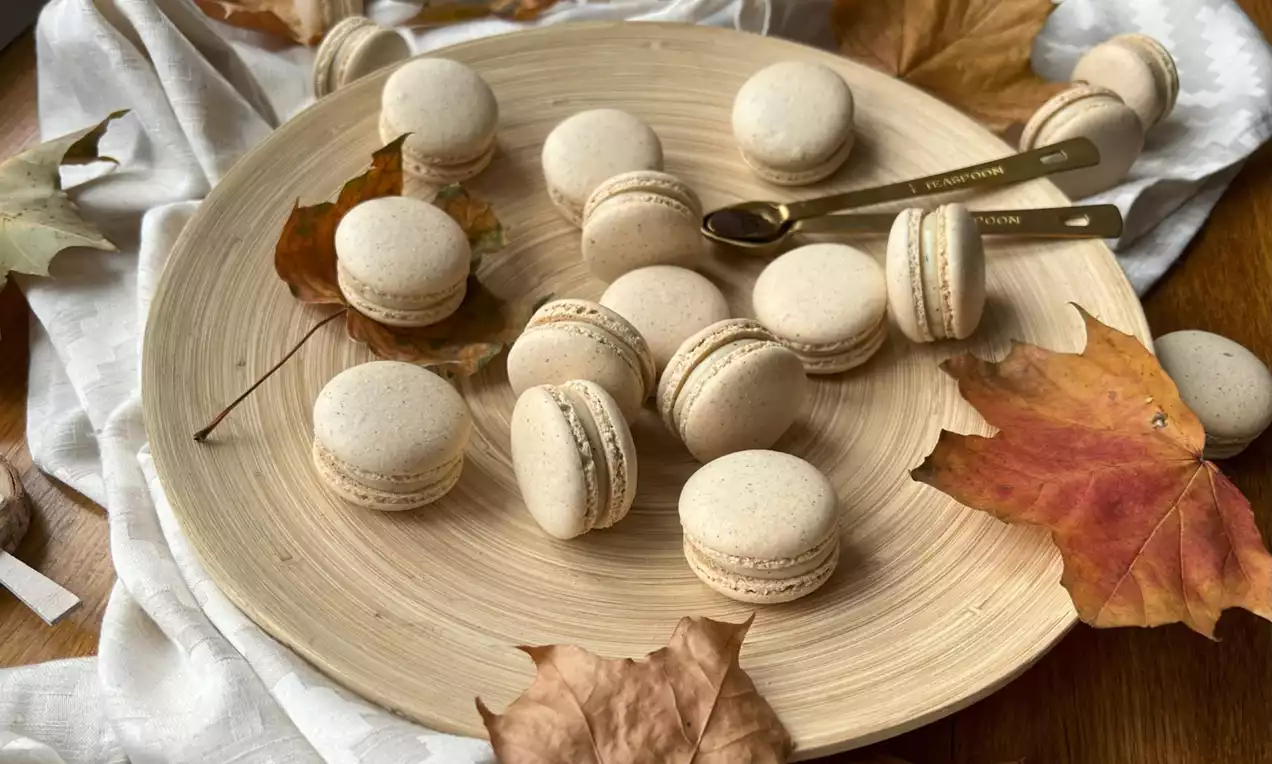 Vanilkové makronky světle béžové barvy s jemným krémem uspořádané na světlém talíři, doplněné podzimními listy a zlatými lžičkami