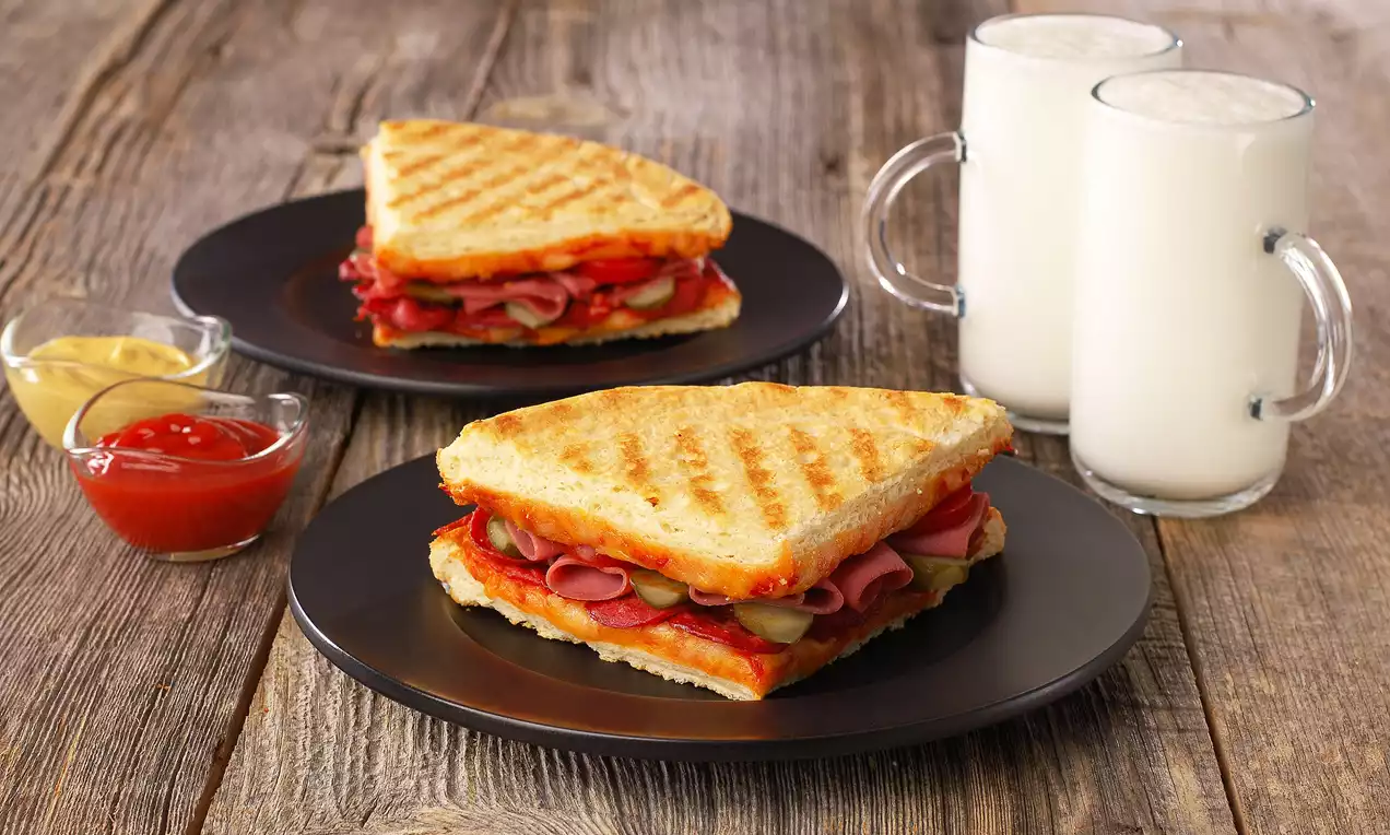 Altın tonlarında kızarmış üçgen tost dilimleri arasında salam, kaşar ve domates yer alırken yanında süt ve soslar sunulmuş siyah tabakta