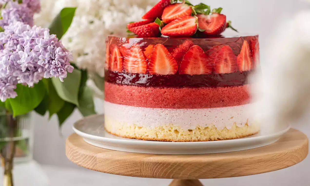 Daudzslāņu torte ar dzeltenu biskvīta pamatni, maigām rozā kārtām un spīdīgu sarkano želeju, dekorēta ar svaigām zemenēm uz koka paliktņa