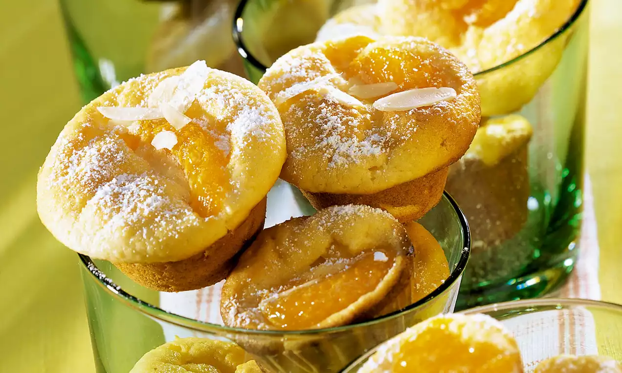Goldgelbe Grieß-Quark-Muffins mit leicht knuspriger Oberfläche, bestäubt mit Puderzucker und Mandelblättchen, angerichtet in grünen Gläsern