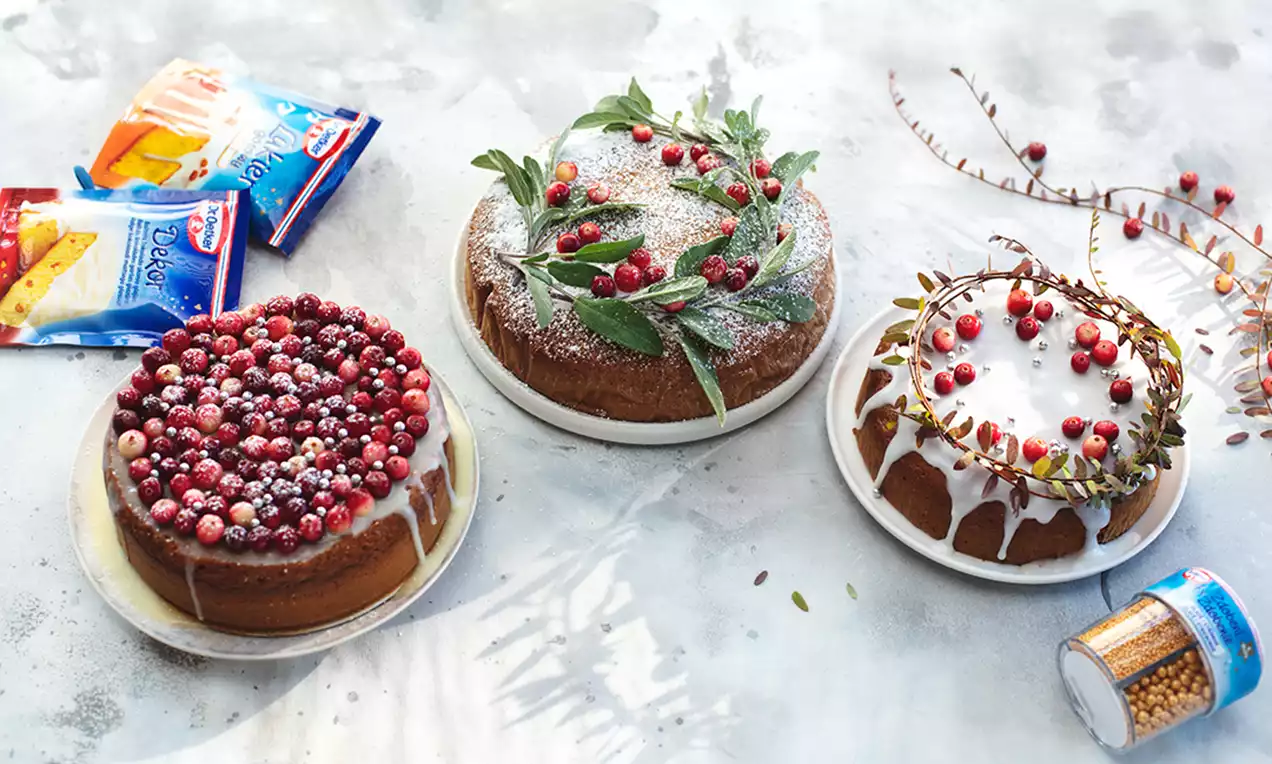 Uz baltas galda virsmas novietotas piparkūkas ar matētu glazūru, blakus čiekuri, dzērveņu zariņi un neliels zilā iepakojumā esošs dekors