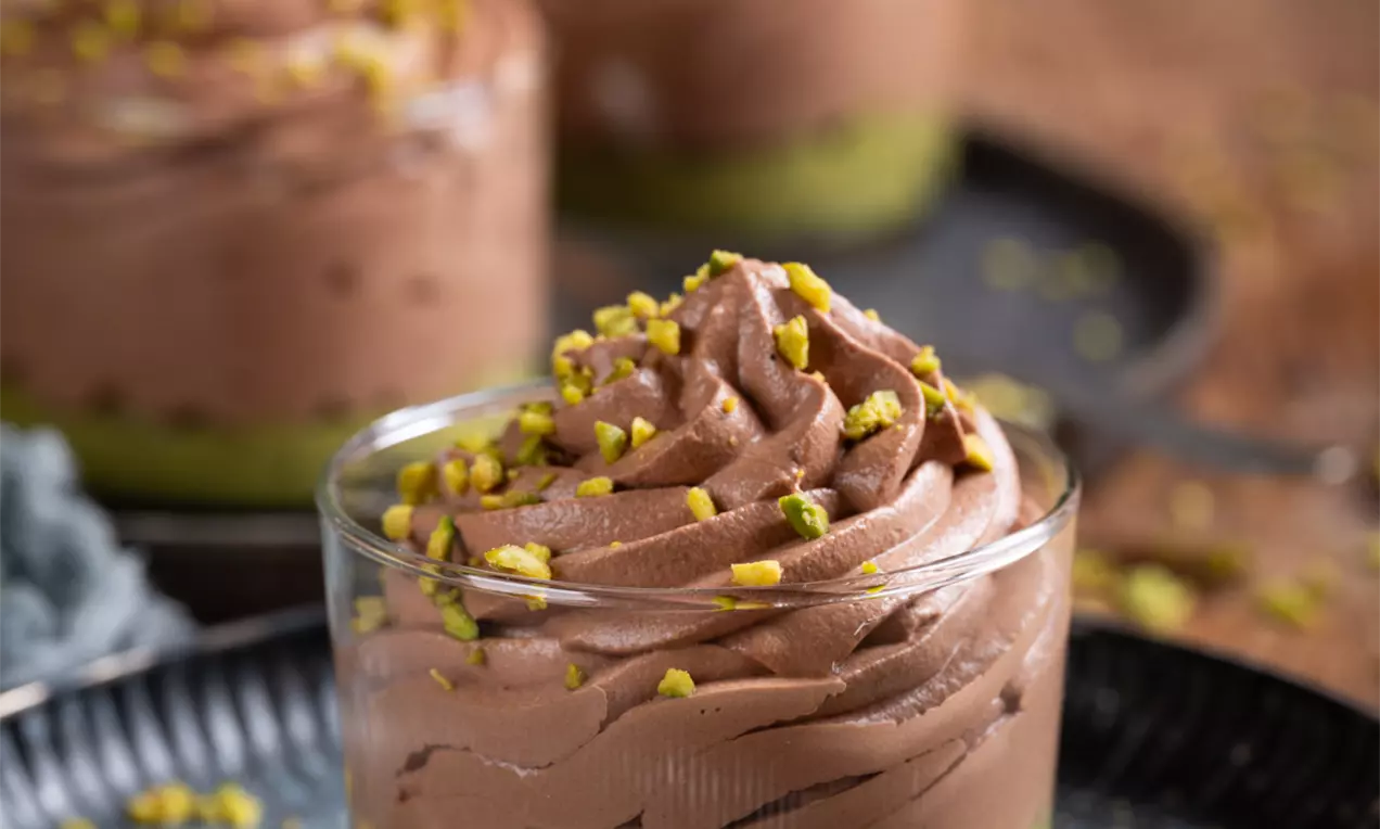 Čokoladna krema svilene teksture složena u vrtložni vrh, posuta nasjeckanim pistaćima na zelenkastom podlošku u staklenoj čaši