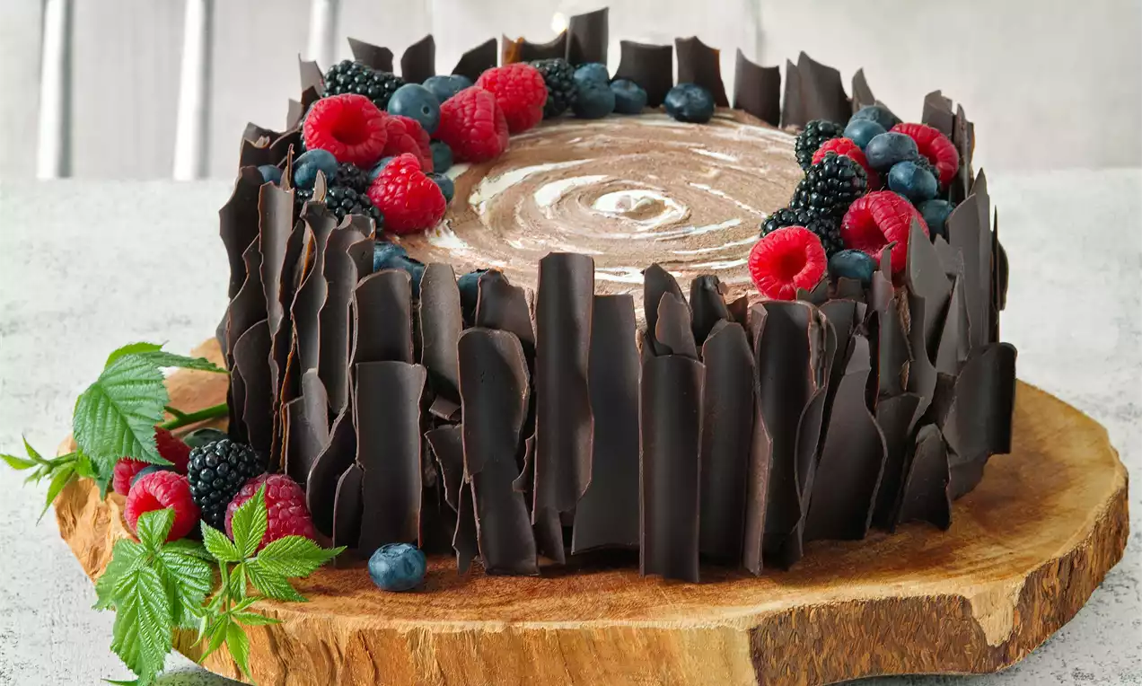 Kakaový dort s kruhovým vírem na krémovém povrchu, lemovaný stočenými čokoládovými plátky, zdobený malinami, borůvkami a lístky na dřevěném podnosu