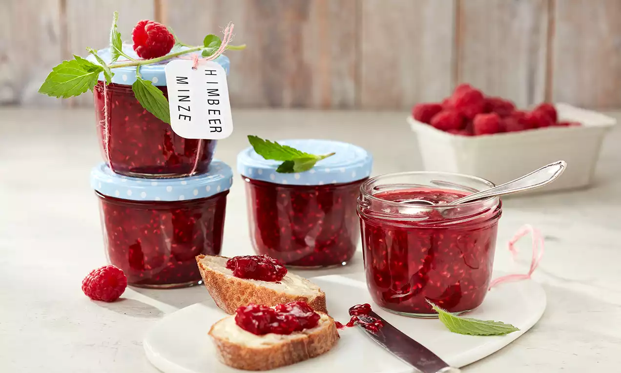 Himbeerrote Konfitüre mit feiner Textur in Gläsern, dekoriert mit Minzblättern, daneben angestrichene Brotscheiben auf weißem Teller