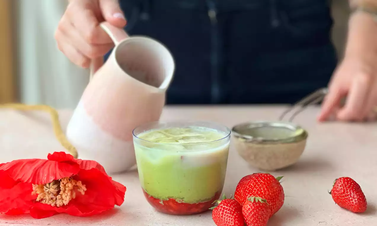 Ein gläsernes Getränk mit grüner Matcha-Schicht, heller Milchschicht und roten Erdbeeren auf hellen Tisch mit Erdbeeren und roter Blüte drapiert