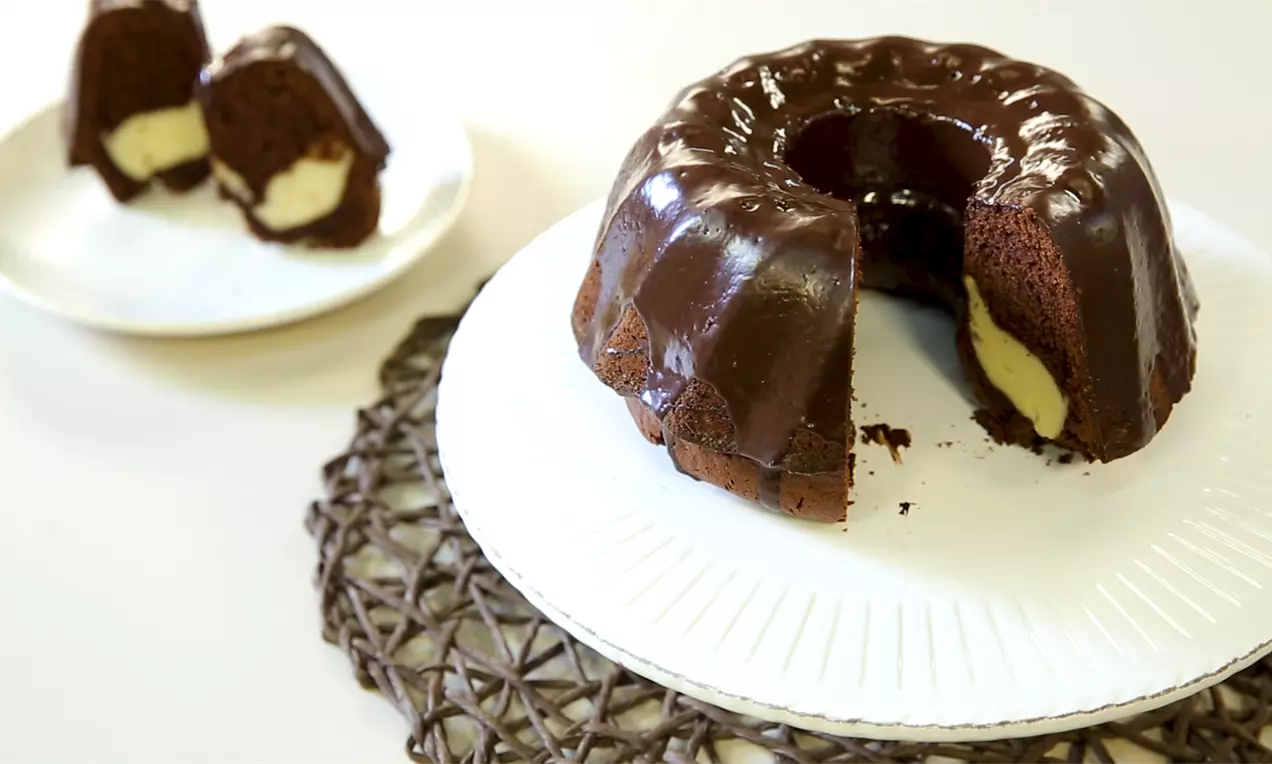 Кръгъл шоколадов кекс с гланцирана глазура и кремава плънка, поднесен върху бяла чиния с разрез, разкриващ вътрешността