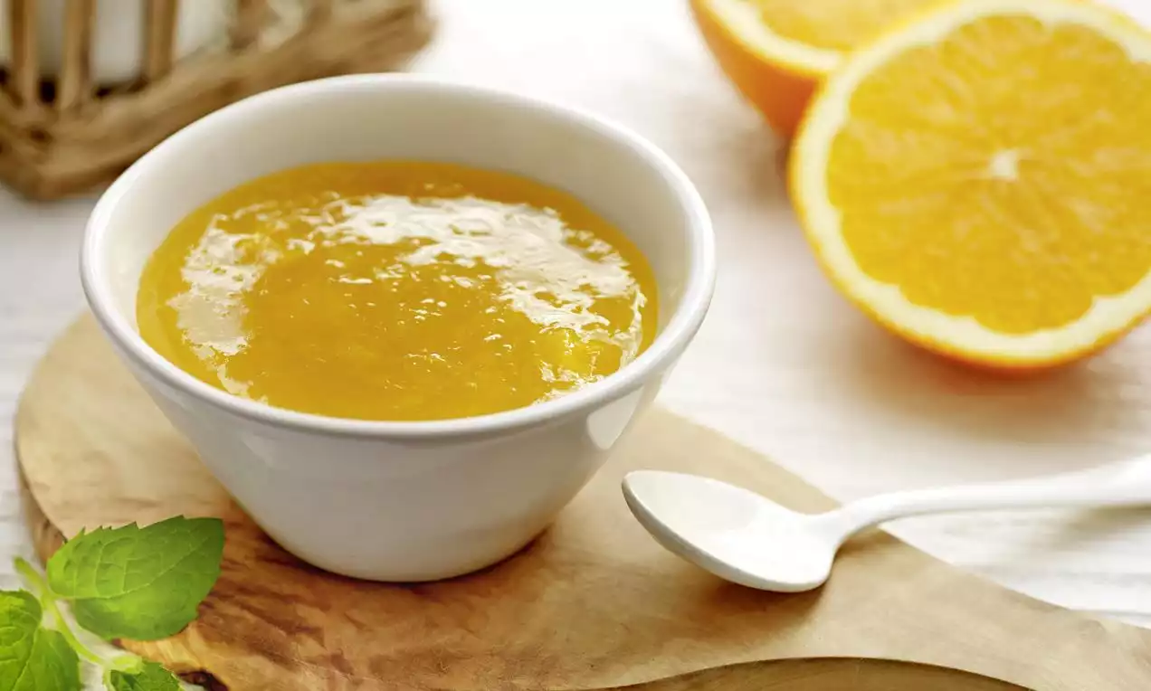 Crema arancione lucida e densa in una ciotola bianca, accompagnata da fette di arancia brillante e un rametto di menta su un tagliere di legno