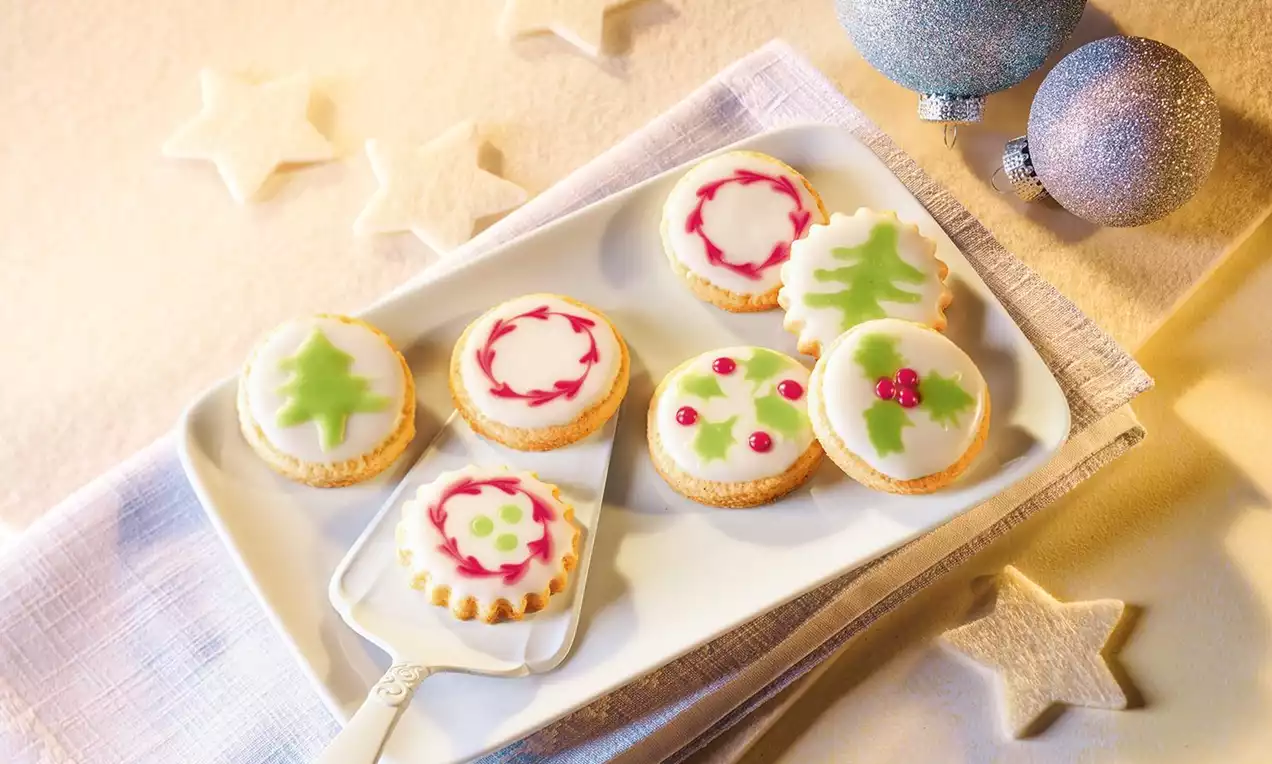 Ronde kerstkoekjes met witte icing en decoraties van groene bomen, rode besjes en sierlijke patronen op een wit bord met kerstversiering