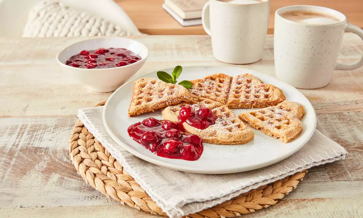 Goldbraune Dinkelwaffeln in Herzform mit Puderzucker und rotem Fruchtkompott auf weißem Teller, dekoriert mit Minze, auf Holzunterlage 