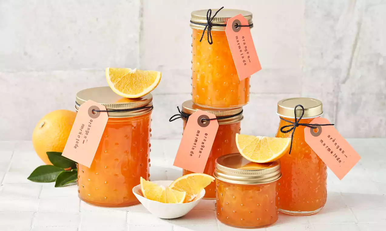 Orangenmarmelade in einem offenen Glas mit glänzender, goldorangener Masse, Löffel auf rustikalem Holzbrett und Orangenscheiben