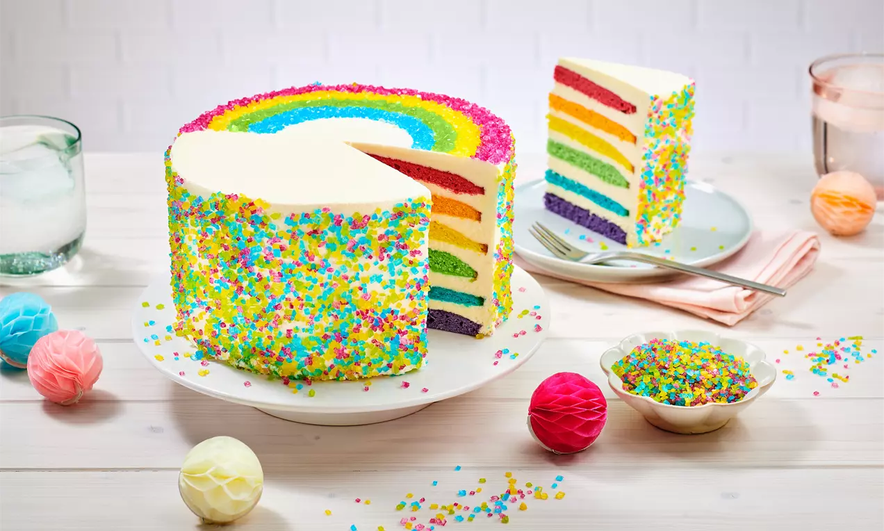 Regenbogentorte mit bunten Schichten und weißer Creme, außen dekoriert mit bunten Zuckerstreuseln, serviert auf weißem Teller mit Beilagen