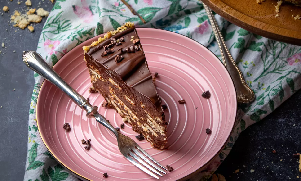 Триъгълно парче торта с плътен тъмнокафяв шоколадов крем и бисквитени парченца, поставено върху розова чиния с разпръснати какаови зърна