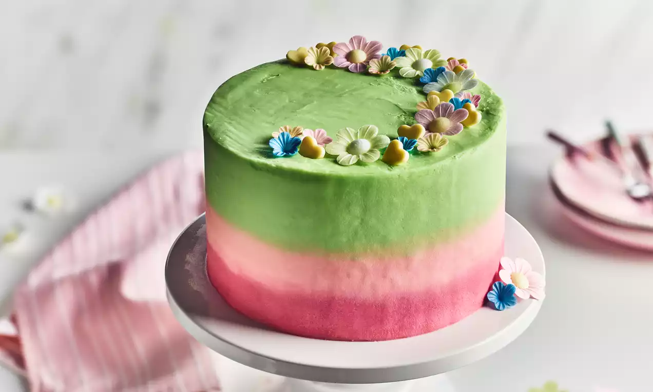Floral ombre cake with smooth green-to-pink gradient frosting, topped with pastel sugar flowers, displayed on a white pedestal stand