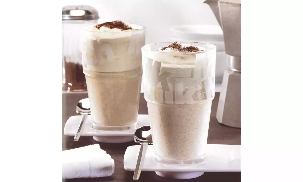 Deux verres transparents remplis de latte macchiato en couches crémeuses, surmontés de mousse blanche et saupoudrés de cacao, posés sur des soucoupes blanches avec une cuillère à côté.