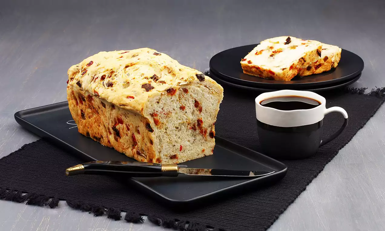 Kuru domates parçalarıyla zenginleştirilmiş somun ekmek, siyah tabakta dilimlenmiş, yanında siyah fincanda kahveyle sunulmuş