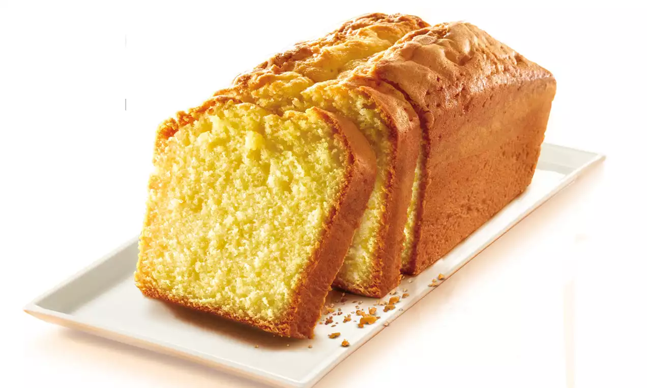 Luchtige goudgele cake met een glanzende bovenkant, in dikke plakken gesneden en liggend op een langwerpig wit bord met kruimels ernaast