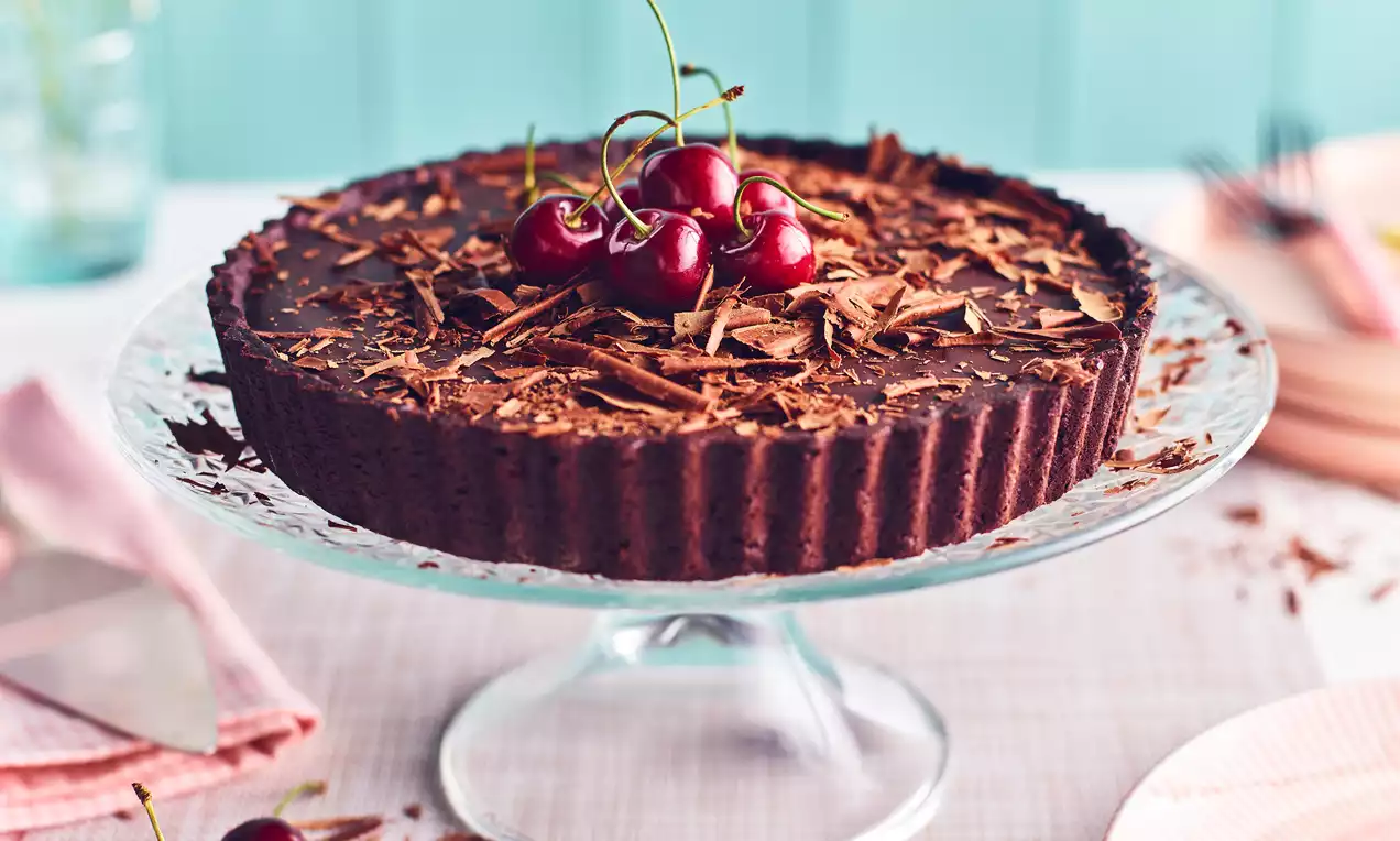 Extra dark chocolate tart with a ridged edge topped with glossy red cherries and dark chocolate shavings on a glass cake stand