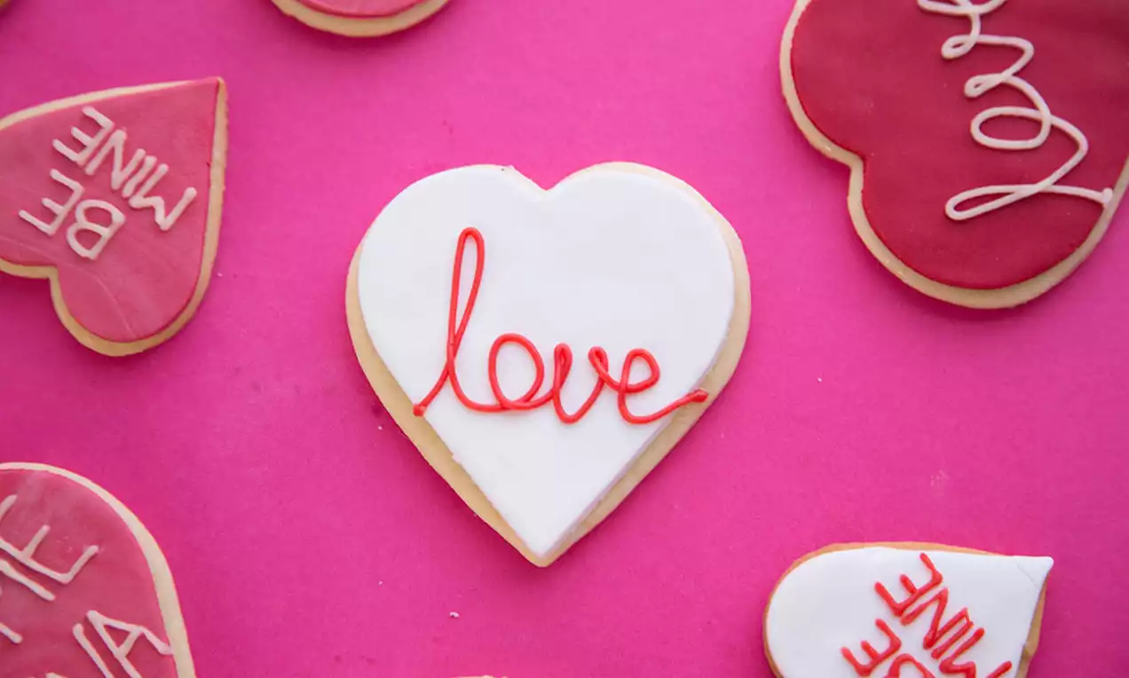 Biscuit în formă de inimă cu glazură albă lucioasă și inscripție roșie subțire, aranjat pe fundal roz alături de alte biscuiți similari