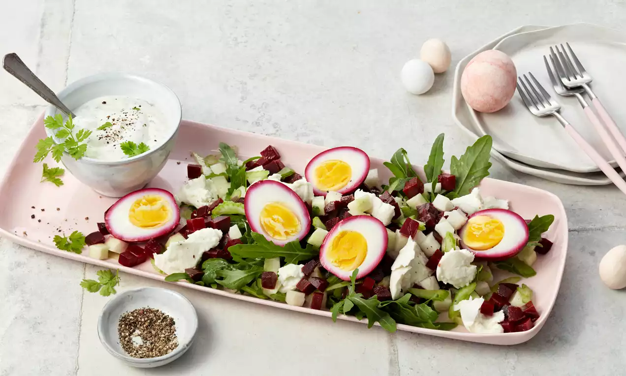 Rote Beete Eier mit gelbem Kern auf Rucola mit weißem Käse, roten Würfeln und Dip in einer Schale auf einem rosa Teller drapiert