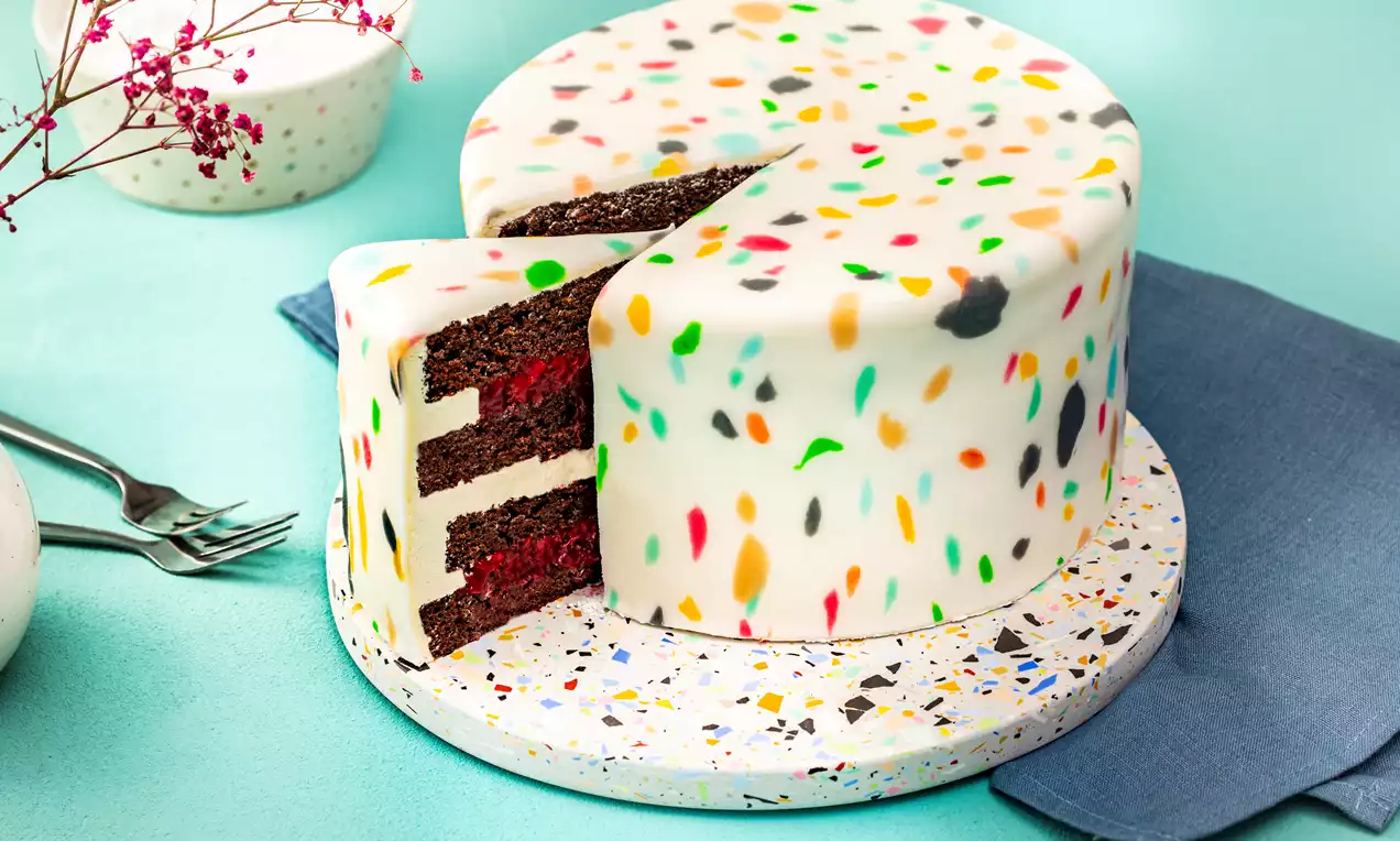 Runder Kuchen mit weißer Glasur und bunten terrazzoartigen Sprenkeln, angeschnitten mit sichtbaren Schichten aus dunklem Teig und roter Füllung