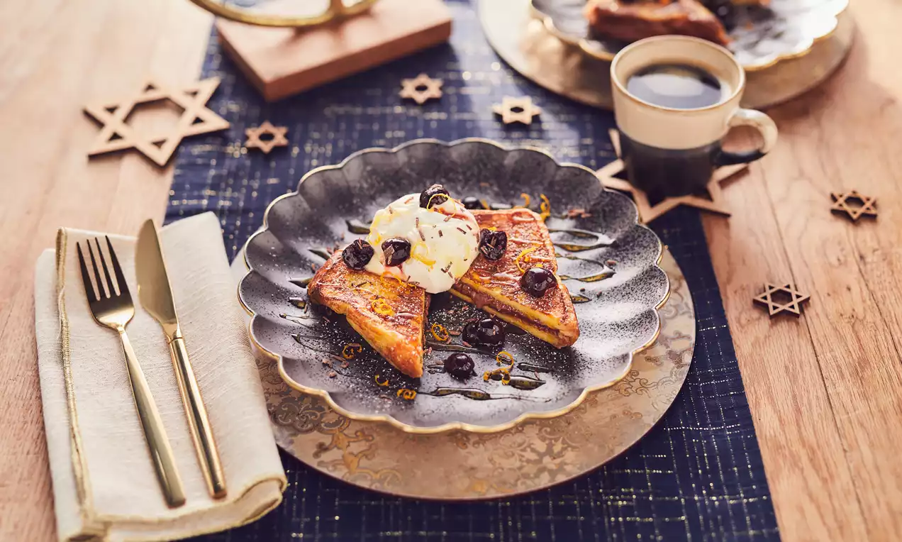 Golden-brown challah bread slices topped with whipped cream, syrup, and dark berries on a scalloped plate with a cup of coffee nearby