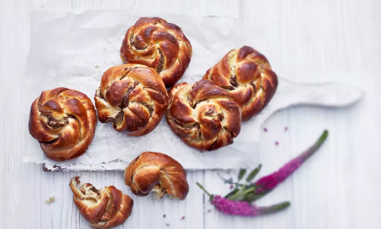 Kiiltävän ruskeiksi paistuneet kardemummakierrepullat pöydällä leivinpaperilla, kierteiden välistä erottuu täytteen pehmeä rakenne