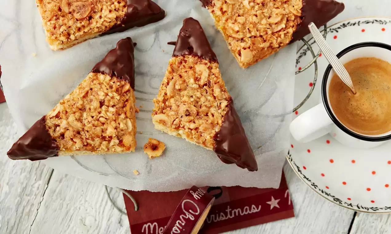 Drei goldbraune Nuss-Ecken mit dunklen Schokoladenspitzen auf weißem Backpapier neben einer weißen Tasse mit Kaffee