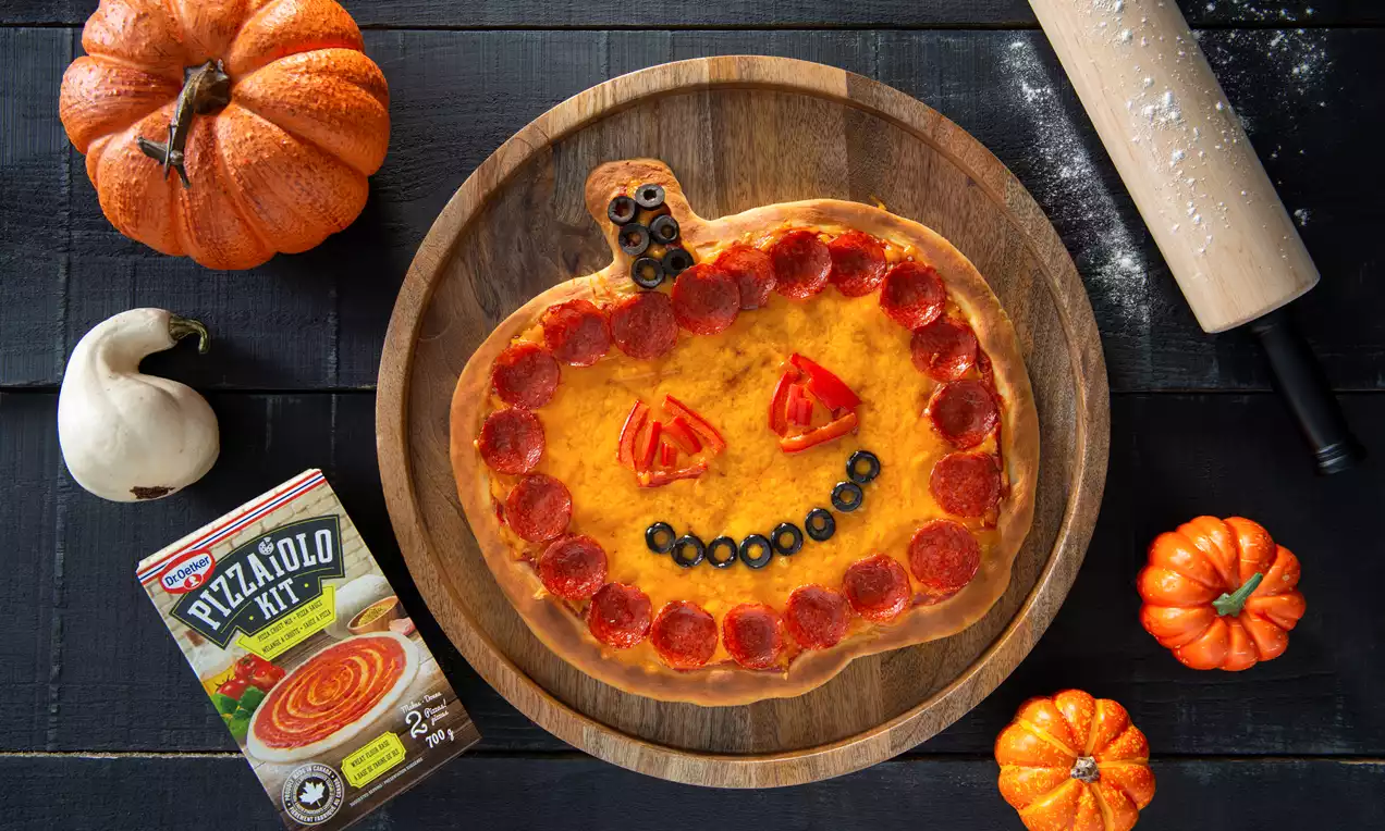 Jack-o’-Lantern-shaped pizza on a wooden plate with orange cheese, pepperoni border, olive smile, and triangle pepper eyes surrounded by pumpkins