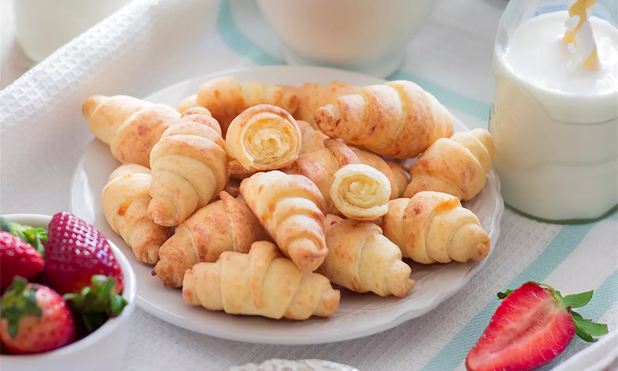 Златисти, леко ронливи кифлички с извита форма, с видими слоеве сирене и кашкавал, подредени върху бяла чиния, до свежа ягода и млечни напитки