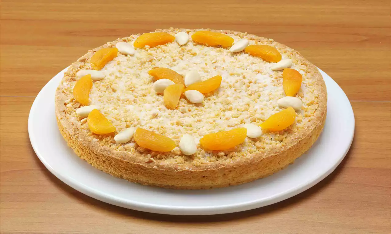 Torta rotonda con base dorata, copertura di briciole friabili, decorata con spicchi di albicocca e mandorle su un piatto bianco
