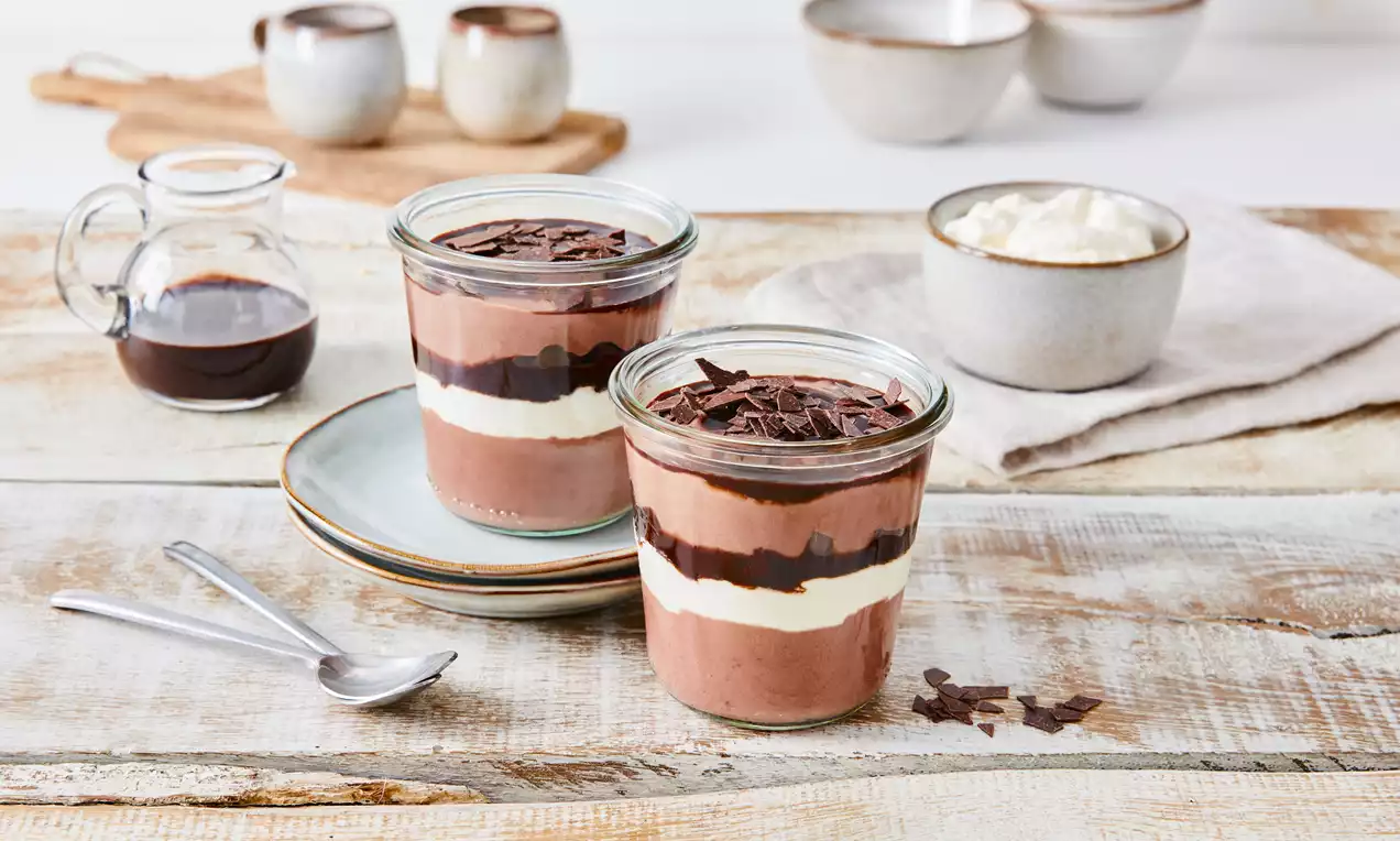 Schokoladendessert im Glas mit Schichten aus heller Sahne, dunklem Schokoladenspiegel und Schokoraspeln auf rustikalem Holztisch