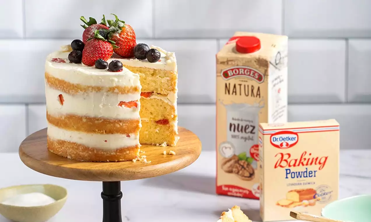 Pastel de capas doradas y esponjosas con relleno blanco, decorado con fresas rojas y arándanos sobre una base de madera