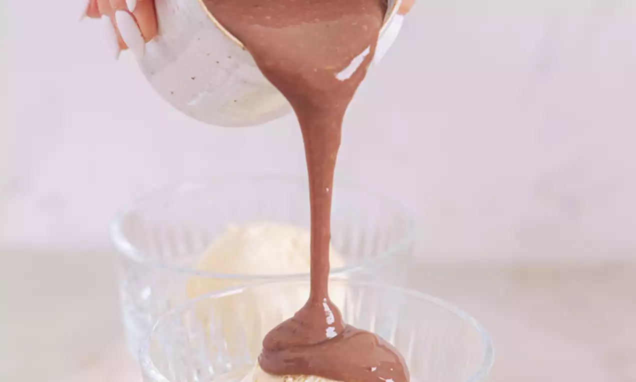 V steklenem kozarcu na podstavku svetlo vanilijev sladoled preliva gosta, temno rjava tekoča čokolada iz bež skodelice