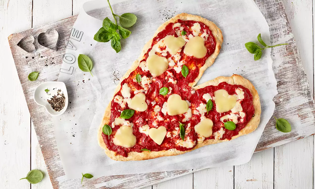Herzförmige Pizza mit roter Tomatensauce, weißen herzförmigen Käsestücken, grünen Basilikumblättern auf Backpapier auf Holzbrett