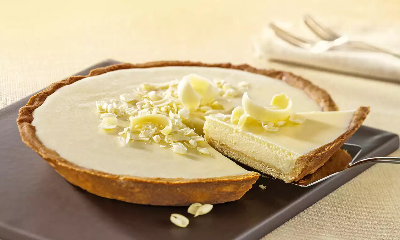 Croccante guscio di frolla marrone con ripieno liscio e chiaro, guarnito con riccioli e scaglie di cioccolato bianco su un piatto scuro