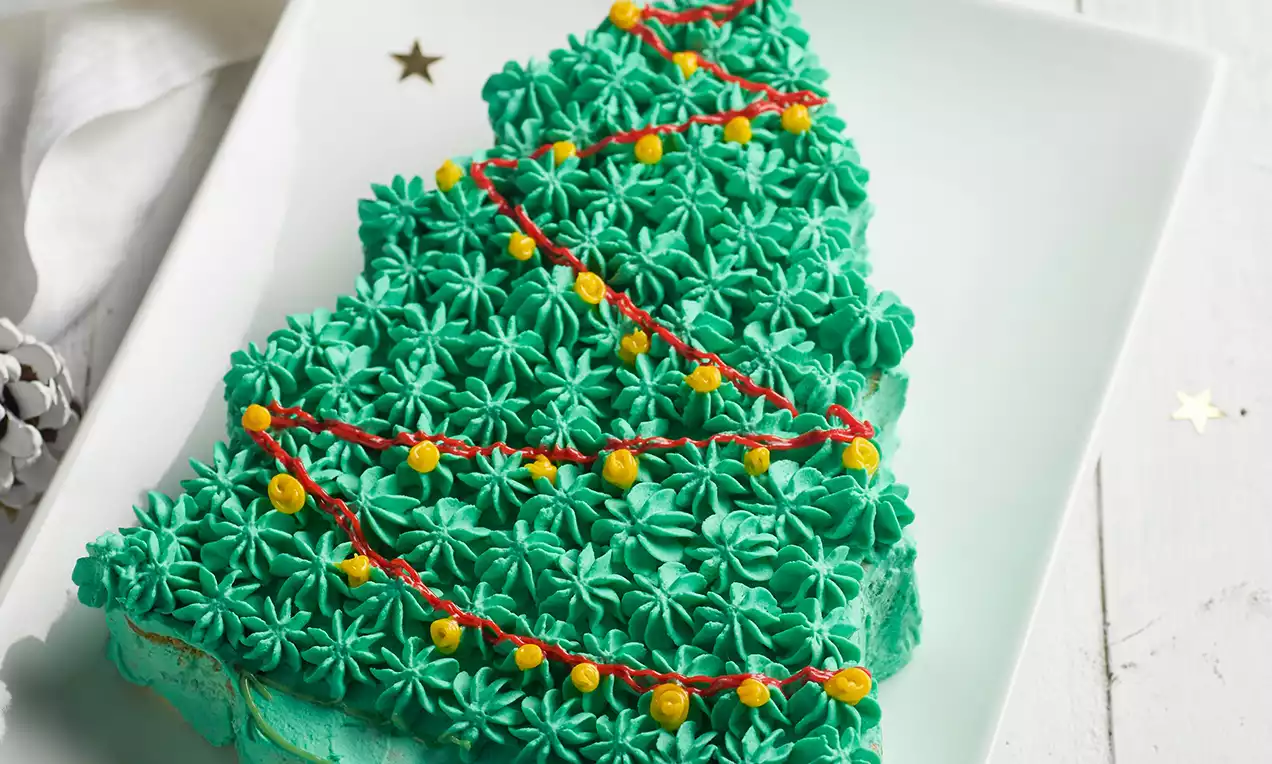 Tarta con forma de árbol decorada con rosetones de crema verde brillante, guirnalda roja y pequeñas esferas amarillas sobre un plato blanco