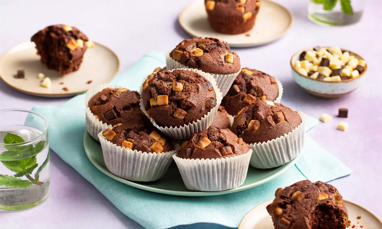 Brioșe ciocolatii dense și umede presărate cu bucăți de ciocolată albă și neagră, așezate pe o farfurie albastră pe un șervet pastel