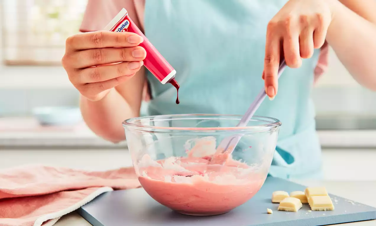 A clear glass bowl with pale pink ganache being mixed, a red gel tube held over it, on a countertop with white chocolate pieces nearby